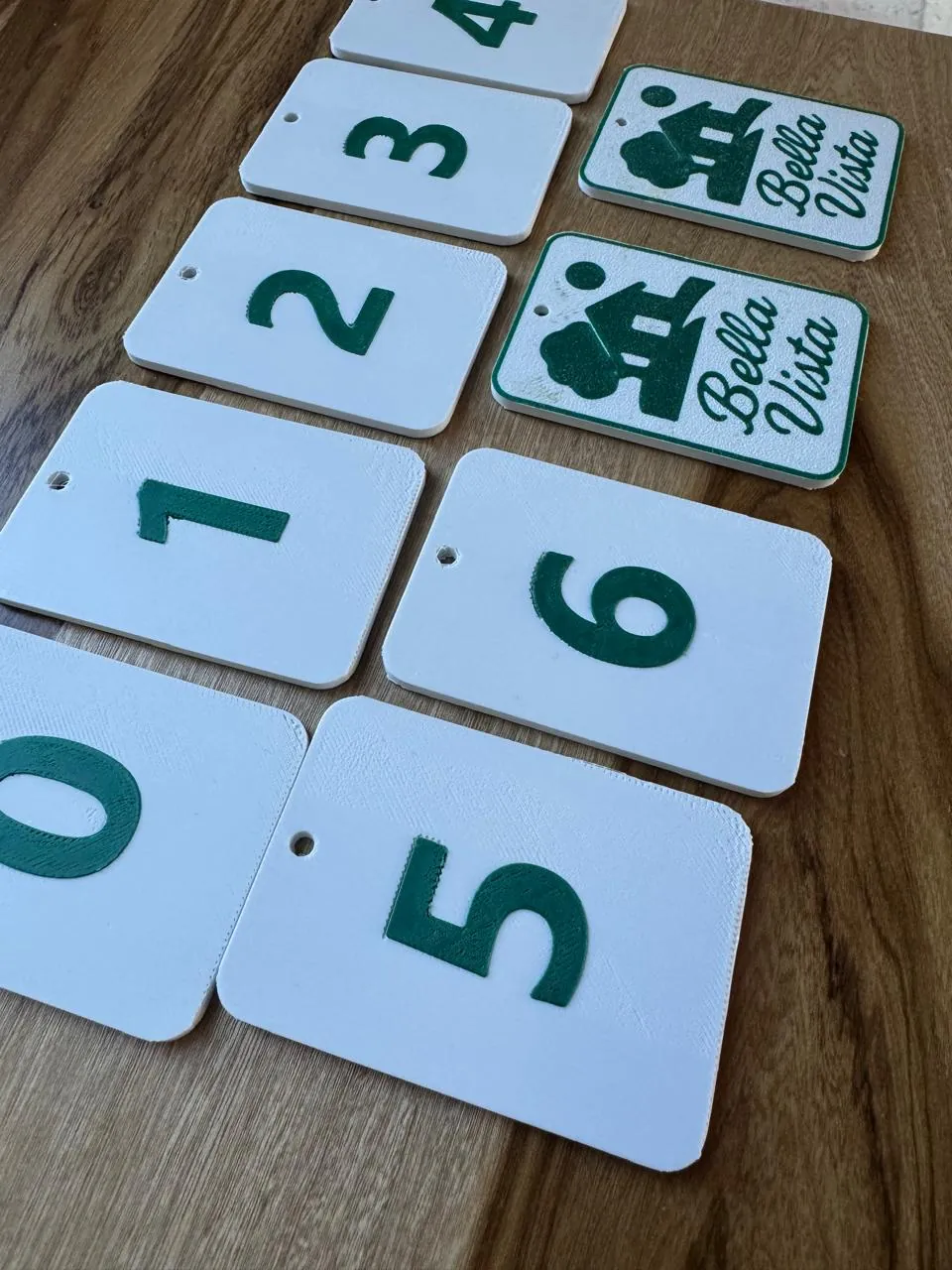 Llaveros rectangulares blancos impresos en 3D, con números verdes del 0 al 6 y dos con el logo de «Bella Vista», sobre una mesa de madera