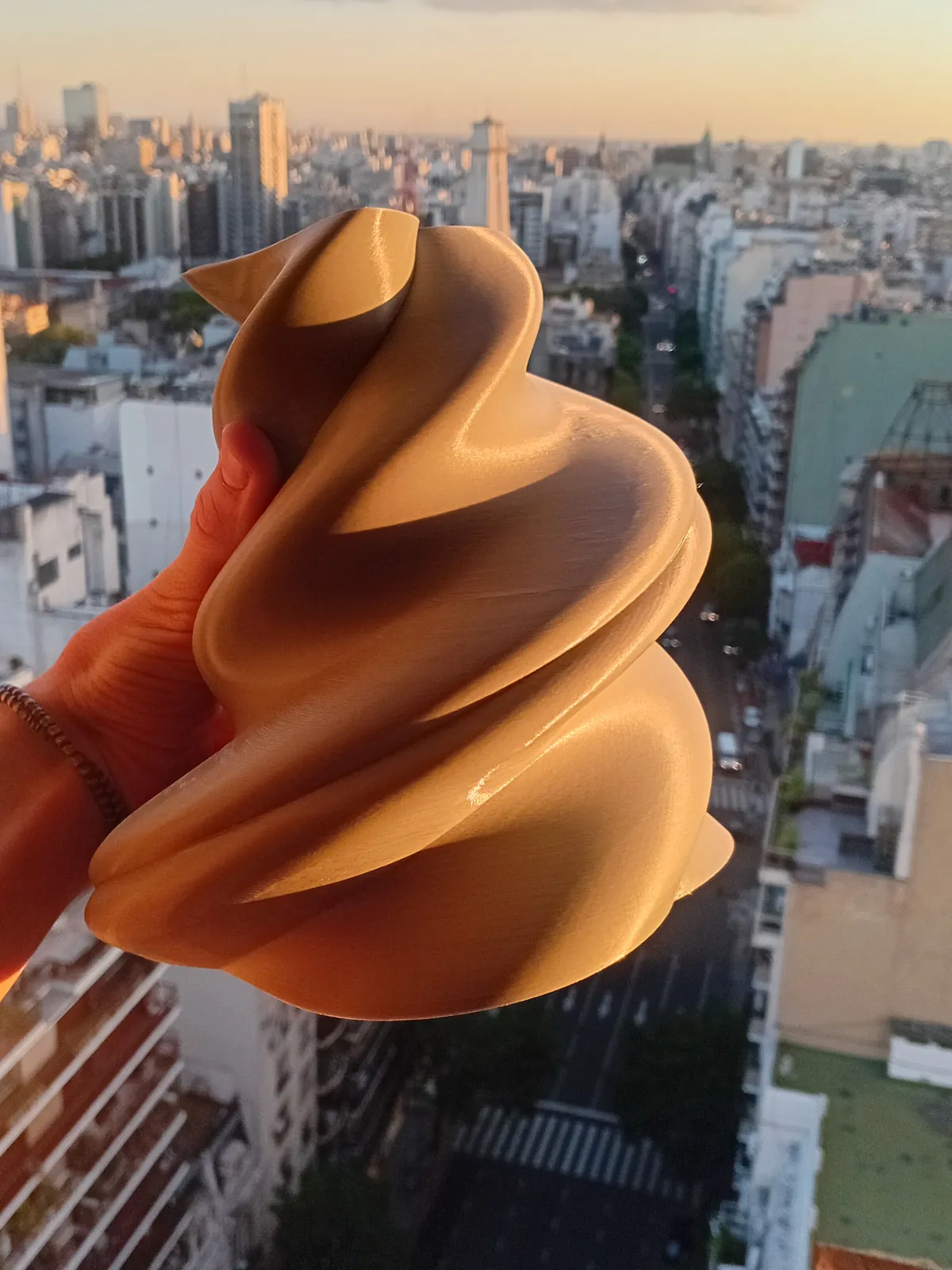 Maqueta de forma orgánica impresa en 3D en tono cobre satinado, sostenida en la mano frente a la ciudad