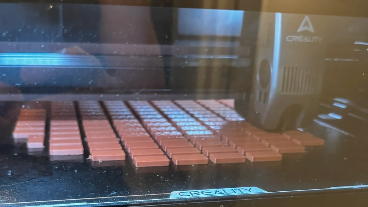 Vista a través de la puerta de la impresora: decenas de piezas rectangulares iguales sobre la cama de impresión