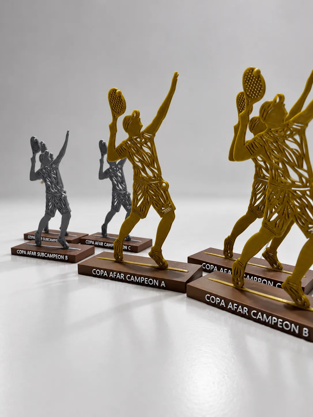 Trofeos de pádel impresos en 3D: siluetas de jugadores en dorado y gris sobre bases con la inscripción de cada categoría